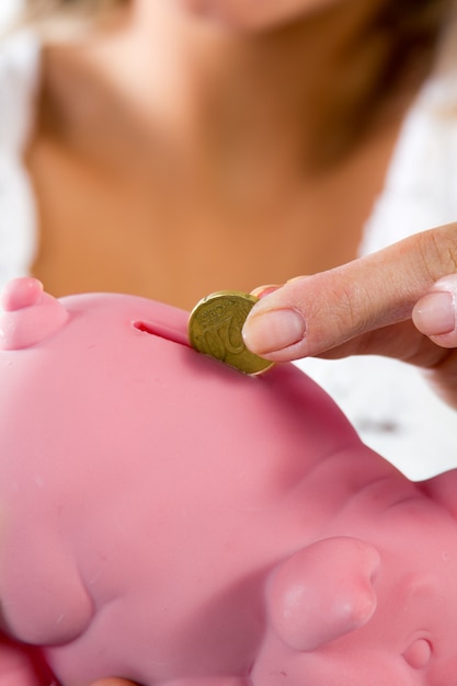 A close-up image of a person's hand carefully placing a coin into a piggy bank, with a soft focus on medical bills in the background, illustrating saving for healthcare expenses.