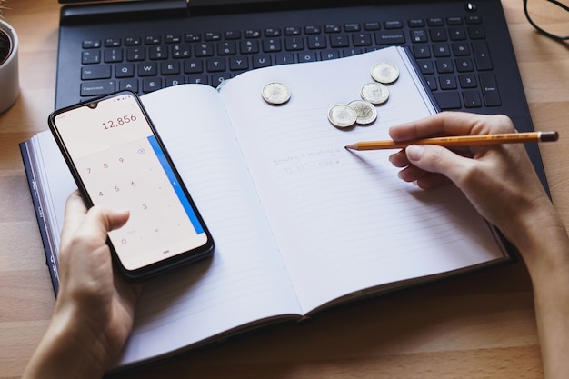 Close up of hands writing in a budget planner, with coins and a calculator nearby, symbolizing meticulous tracking and planning.