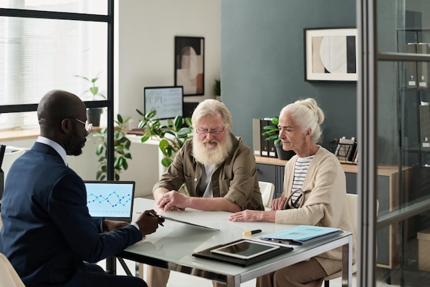 An elderly couple consulting with a financial advisor in a bright and comfortable office setting. The focus is on the advisor explaining healthcare planning options using visual aids like charts and graphs.