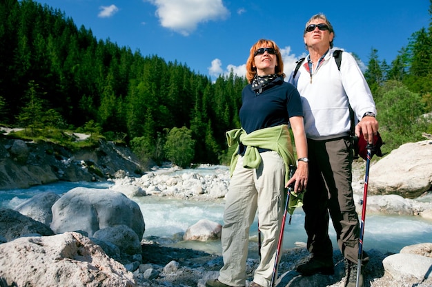 A couple enjoying their retirement, hiking in a scenic mountain area, wearing comfortable outdoor clothing. The image conveys a sense of freedom, adventure, and active living during retirement. The sunlight is warm and inviting.